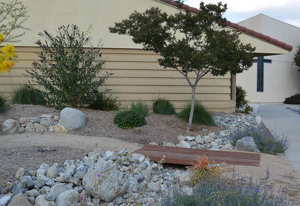 Dry creek bed with boulders, a small wooden bridge, and drought-tolerant planting beside a church building.